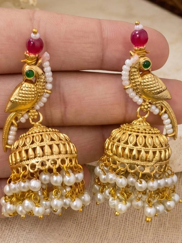 Close-up of gold parrot jhumka earrings with red beads and dangling white pearl drops held in a hand