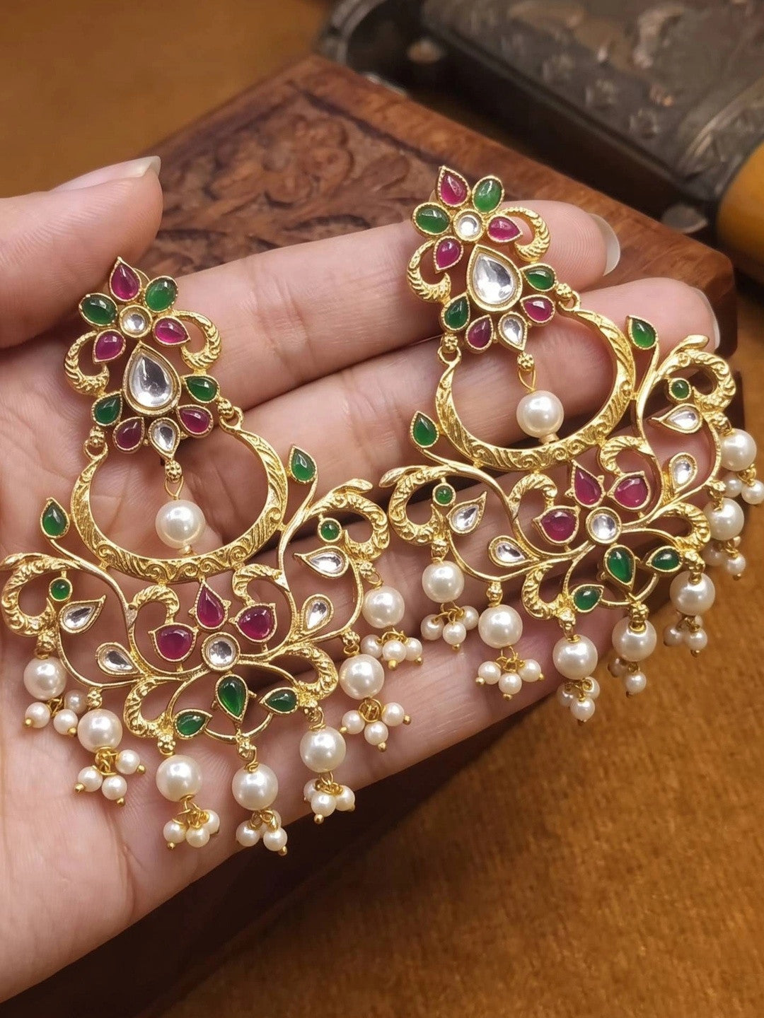 Close-up of gold-plated kundan chandbali earrings held in a palm showing scrollwork, coloured stones and cascading pearl drops