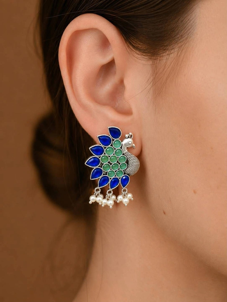 Oxidised silver peacock stud earring worn on ear showing blue and green stones with pearl drops