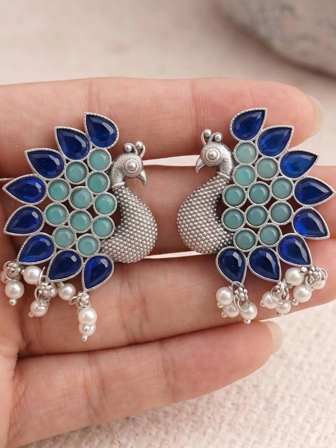 Pair of peacock stud earrings with blue teardrop and mint green stones held in palm