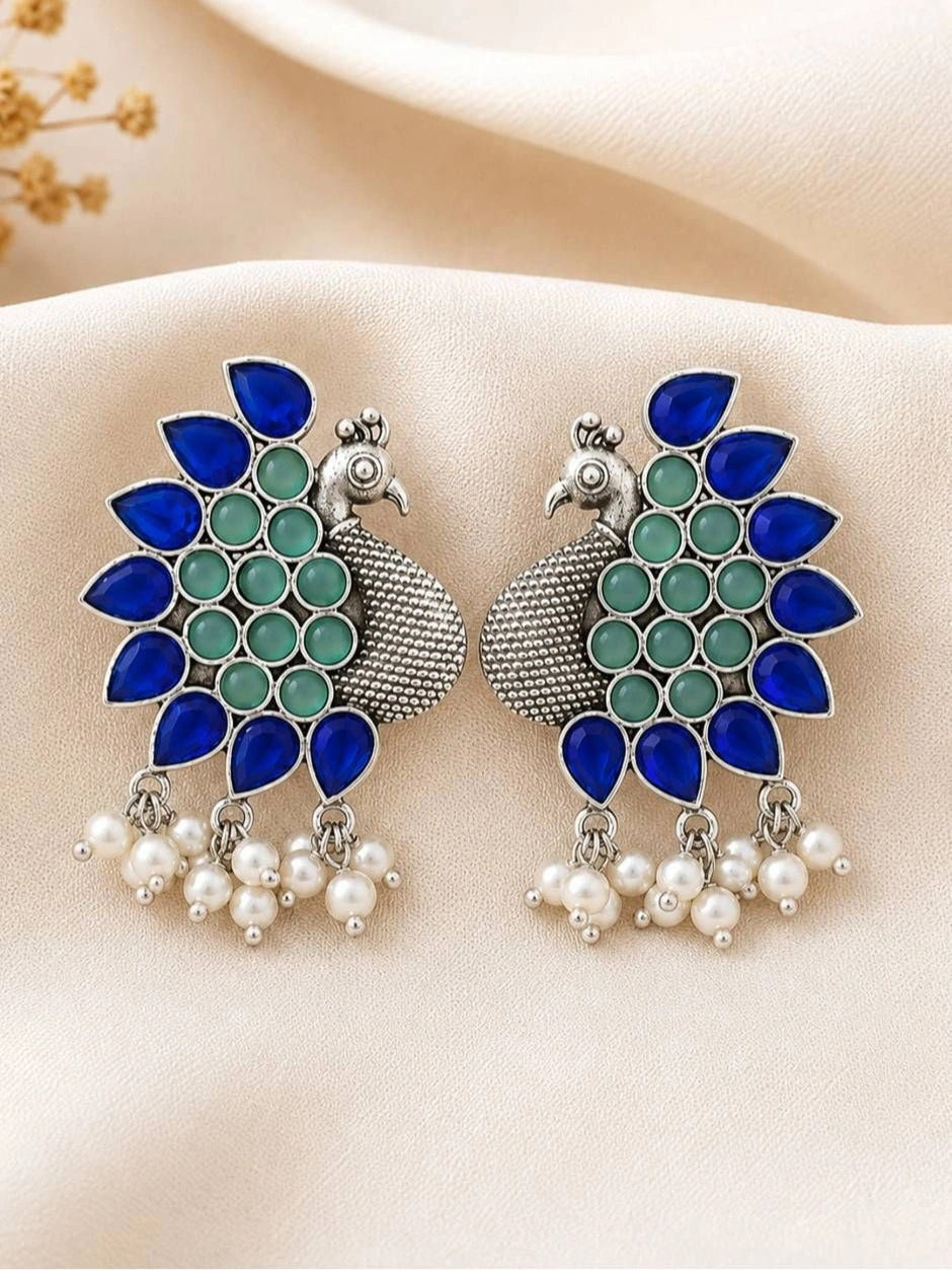 Two oxidised silver peacock earrings with pearl danglers laid flat on white satin