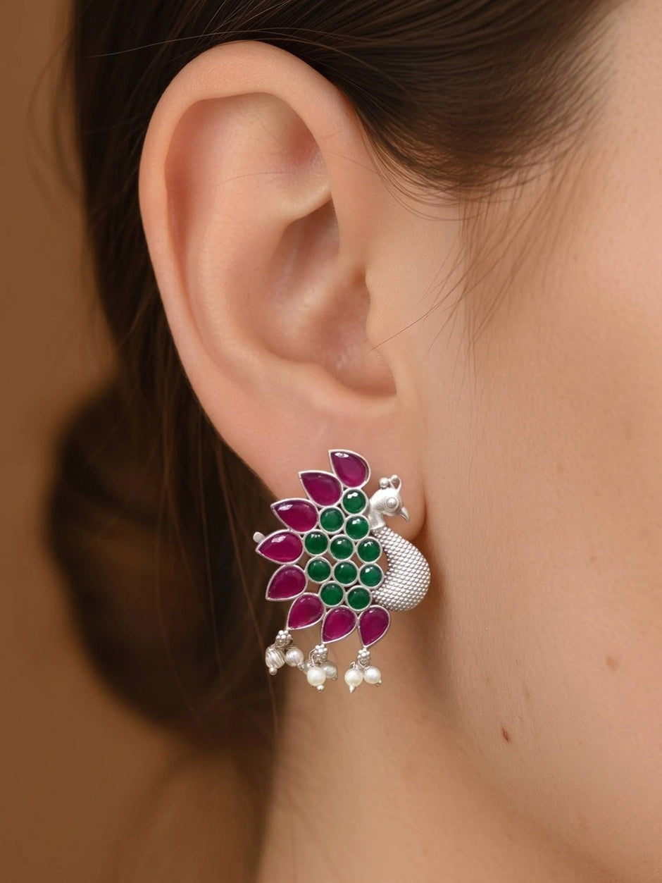 Oxidised silver peacock stud earring worn on a woman's ear, showing magenta and green stones with pearl drops