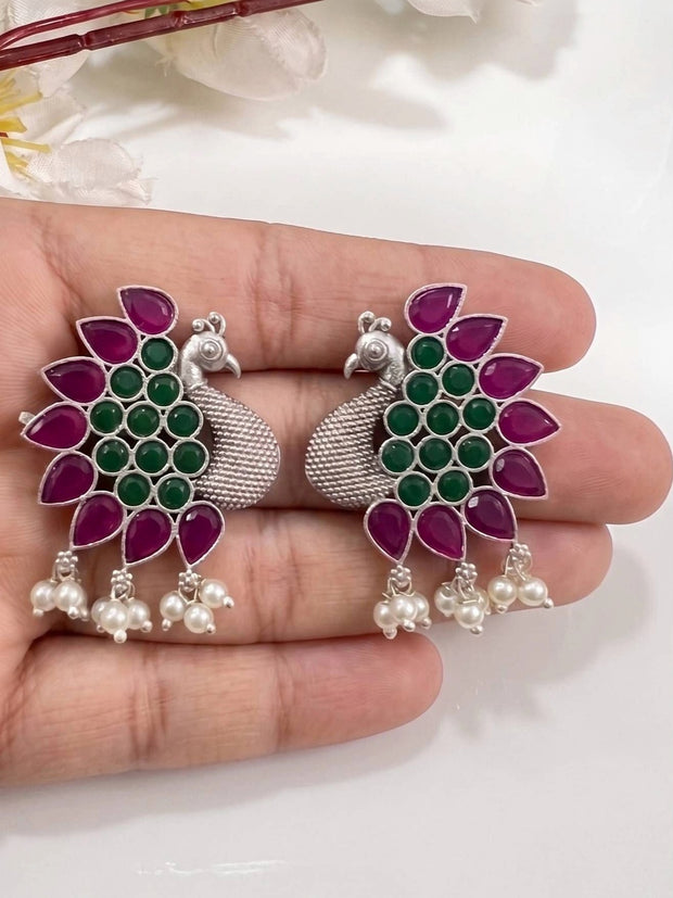 Pair of peacock stud earrings held in an open palm showing full front design with stones and pearl dangles