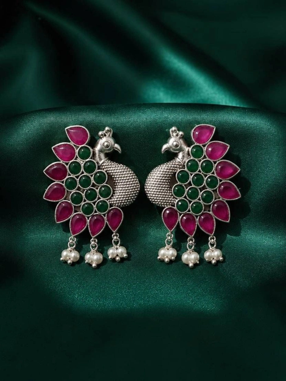Pair of oxidised silver peacock earrings laid flat on green silk fabric showing mirrored peacock silhouettes