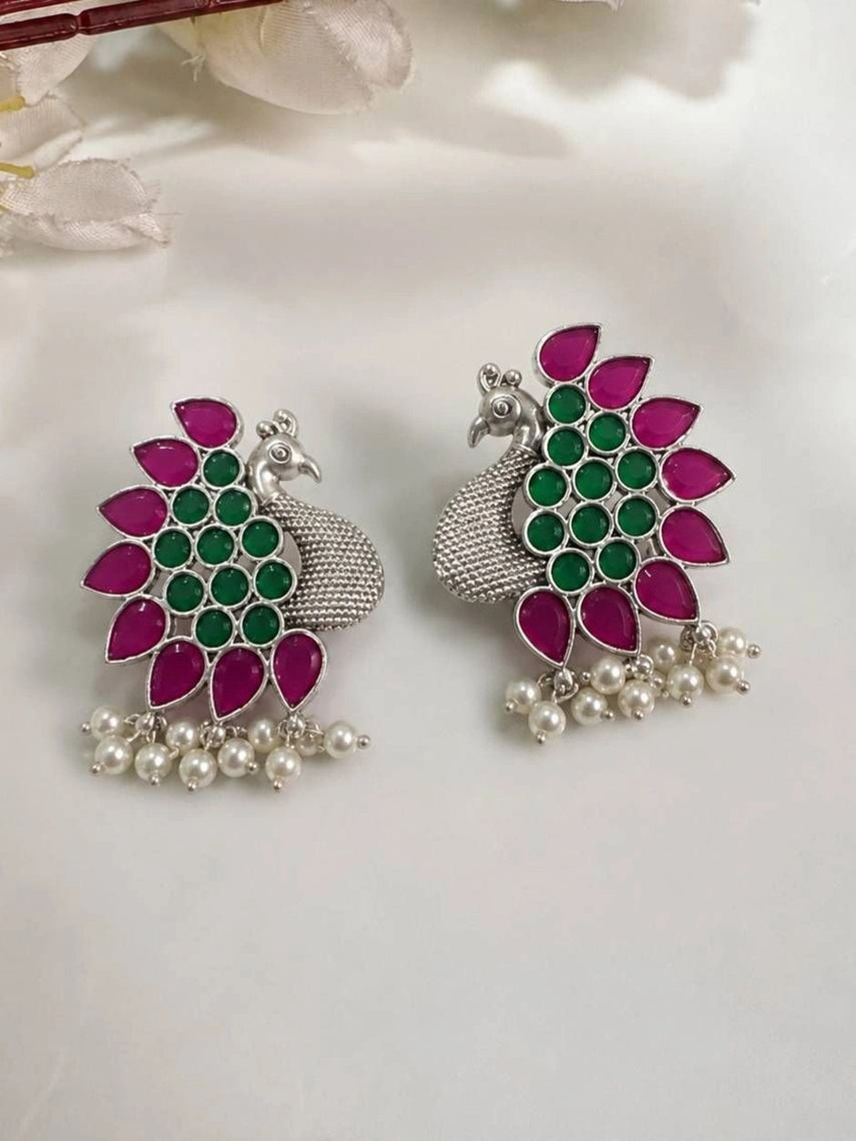 Two peacock stud earrings side by side on white fabric highlighting textured silver body and coloured stone tail