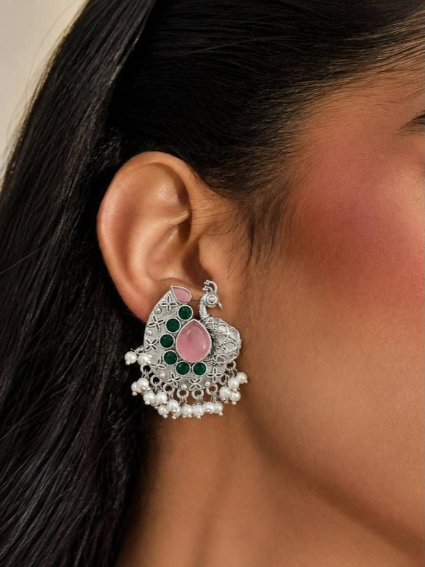 Pair of peacock chandbali earrings held on a palm, showing pink teardrop stones and green cabochons