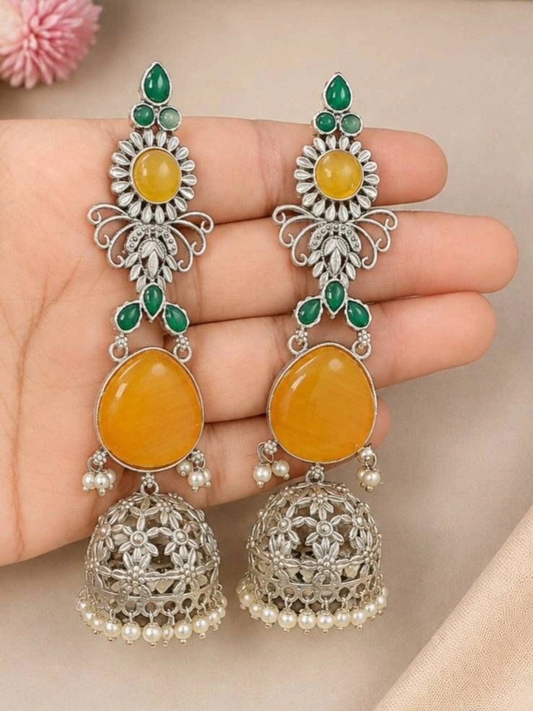 Pair of oxidised silver jhumka earrings with yellow cabochons and green stones resting on an open palm