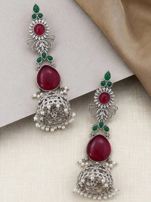 Pair of oxidised silver jhumka earrings with red and green stones laid flat on a cream surface