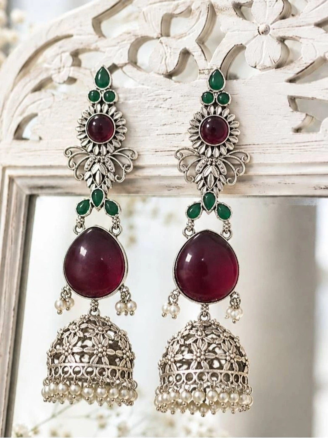 Pair of ruby and green stone jhumka earrings displayed on a white carved wooden frame