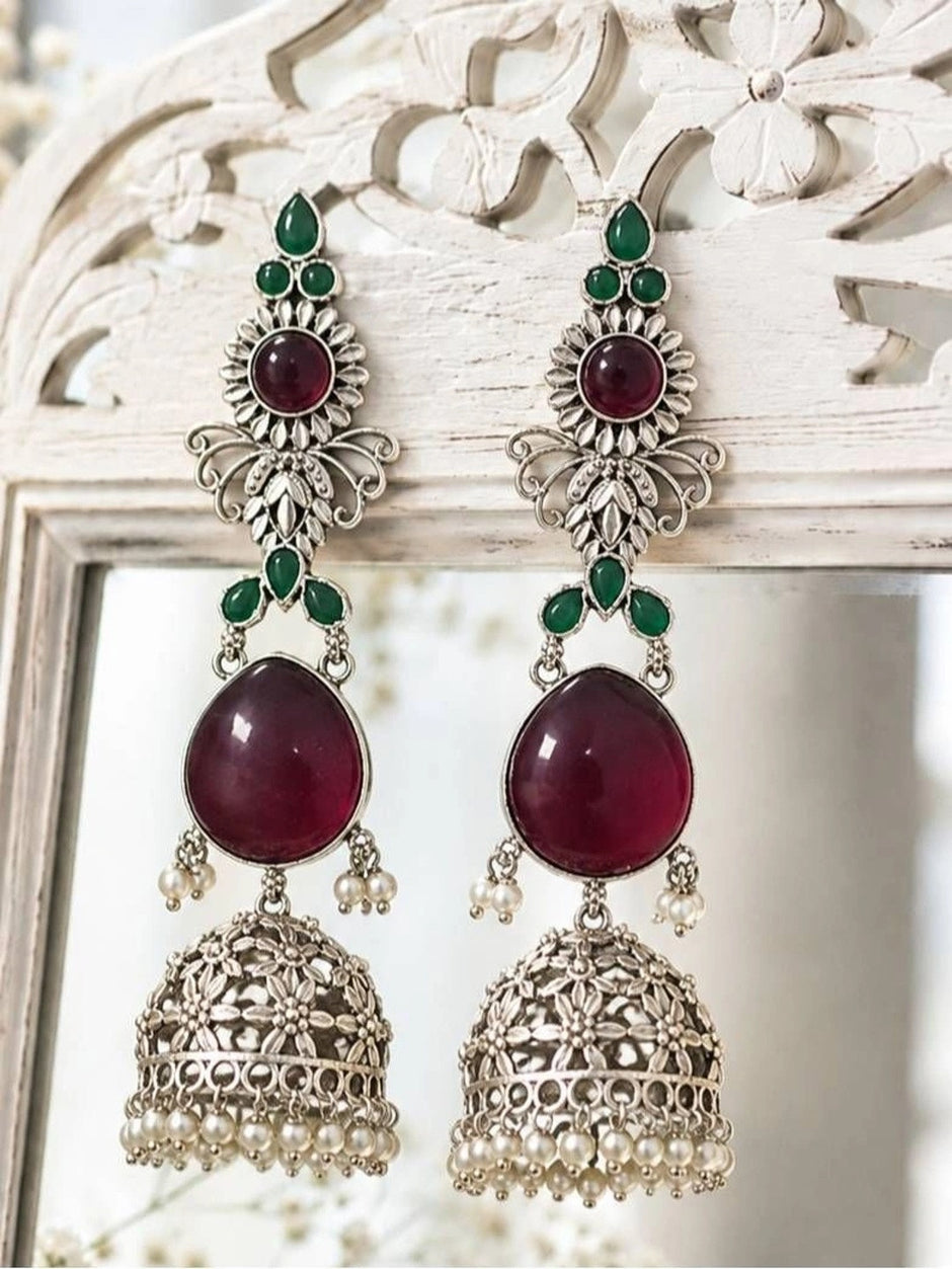 Pair of ruby and green stone jhumka earrings displayed on a white carved wooden frame