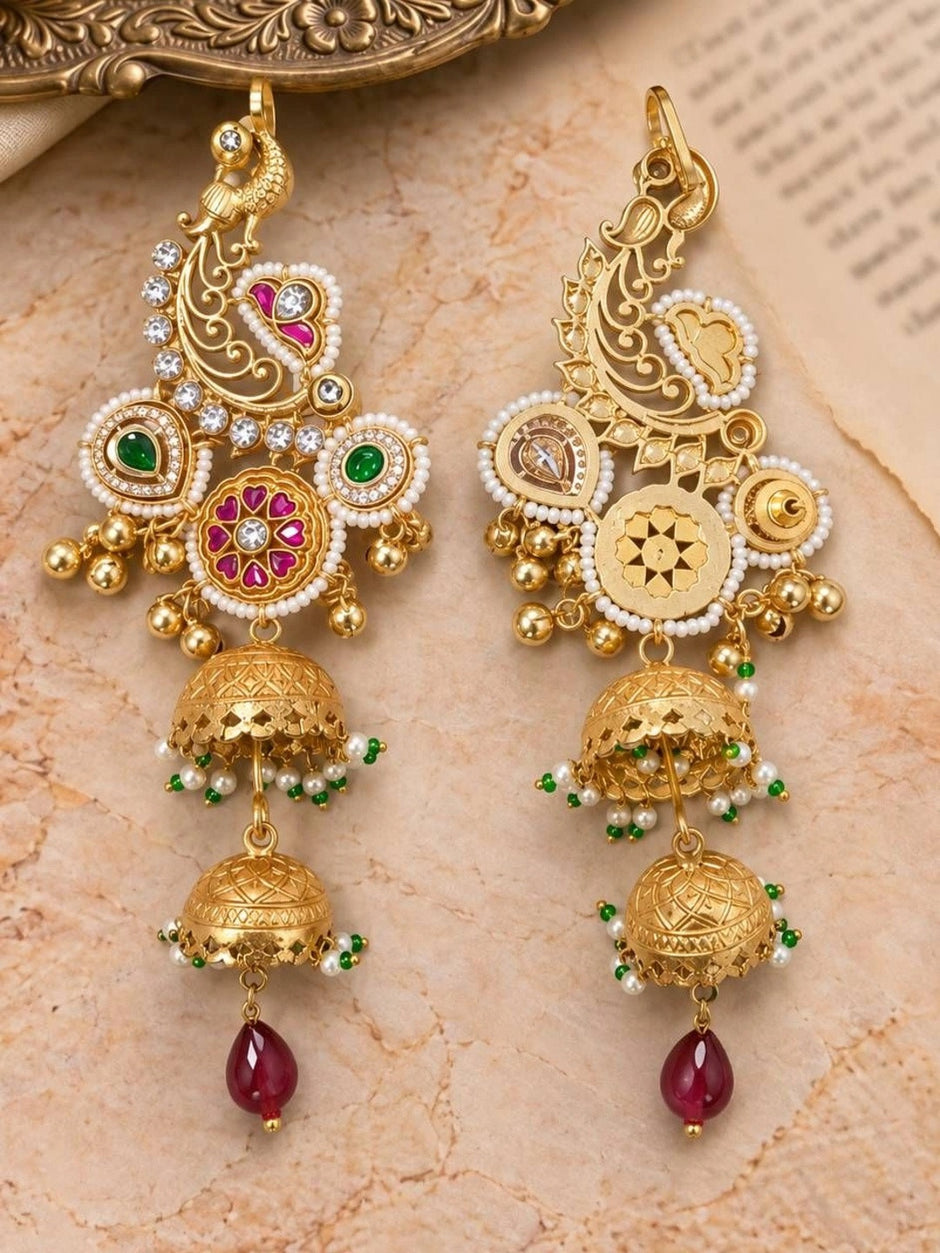 Front view of two gold chandbali jhumka earrings with ruby and emerald accents laid on a marble surface