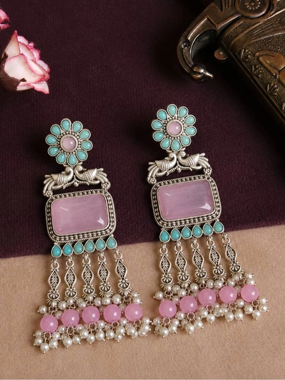 Pair of oxidised silver chandbali earrings with peacock motif and pink pearl cluster tassels on a dark maroon background with roses