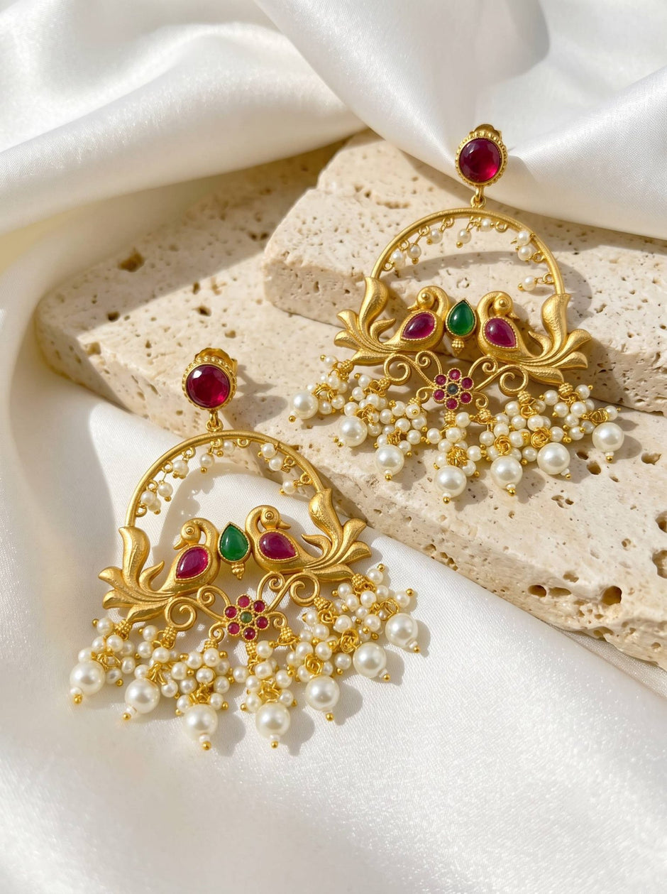 Pair of gold-plated peacock chandbali earrings with red green stones and white pearl drops on white satin and stone surface