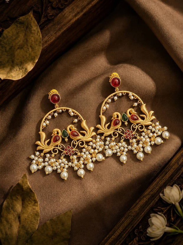 Two gold-plated chandbali earrings with peacock motifs and pearl clusters displayed on brown silk fabric with dried leaves