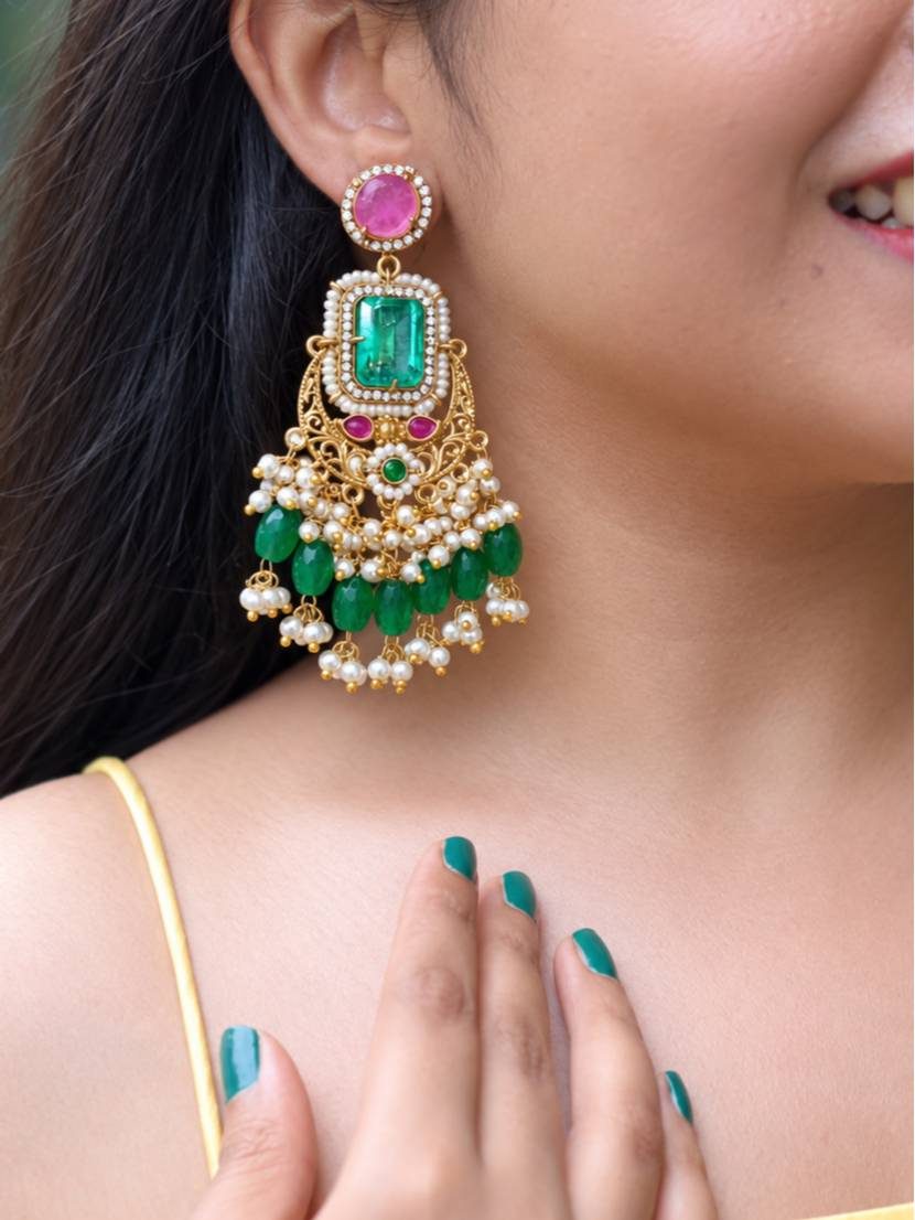 Close-up of a woman wearing a gold-plated emerald green chandbali earring with pearl drop fringe, paired with a yellow outfit and teal nail polish