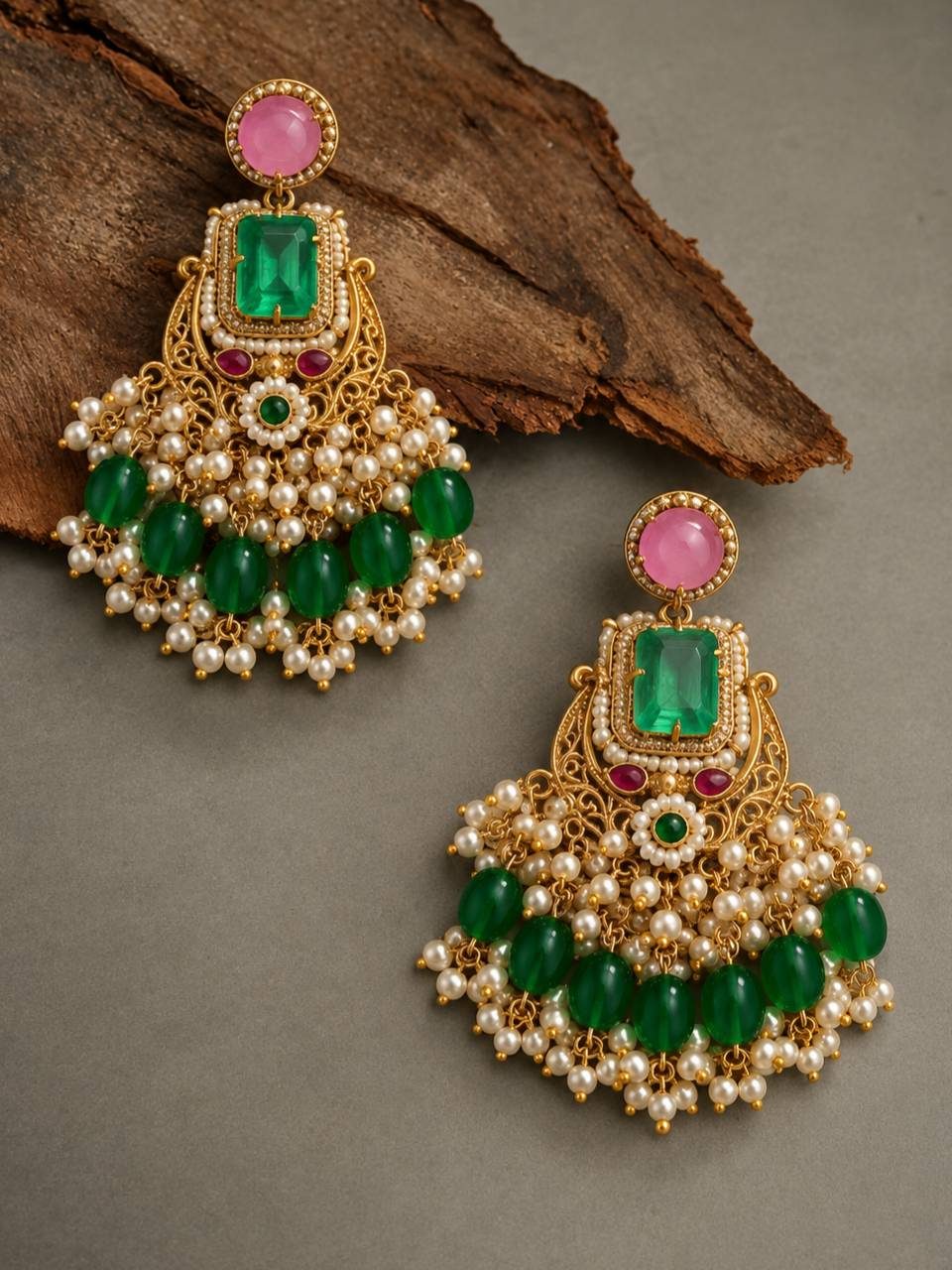 Two gold-plated chandbali earrings with emerald green oval bead drops and seed pearl detailing, laid flat on a grey surface beside a piece of bark