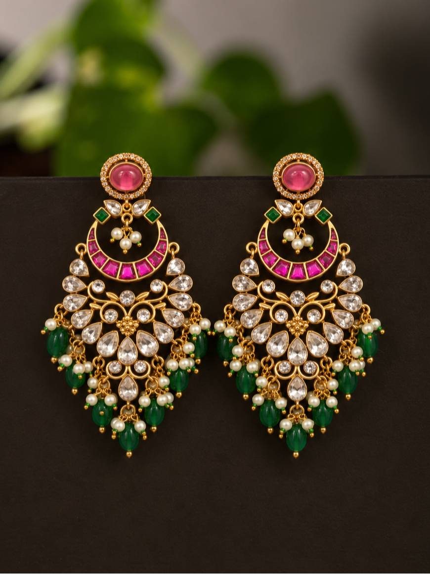 Pair of gold-plated kundan chandbali earrings with pink stone tops, fuchsia crescent arch, white kundan panel, emerald green bead drops and seed pearls displayed on dark background