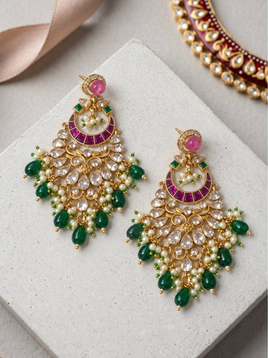 Two gold-plated kundan chandbali earrings laid flat on white stone surface showing front face with pearl clusters and green drop beads