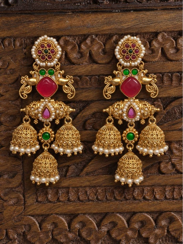 Pair of antique gold-plated triple jhumka earrings with pink stone and pearl drops displayed on carved wooden surface