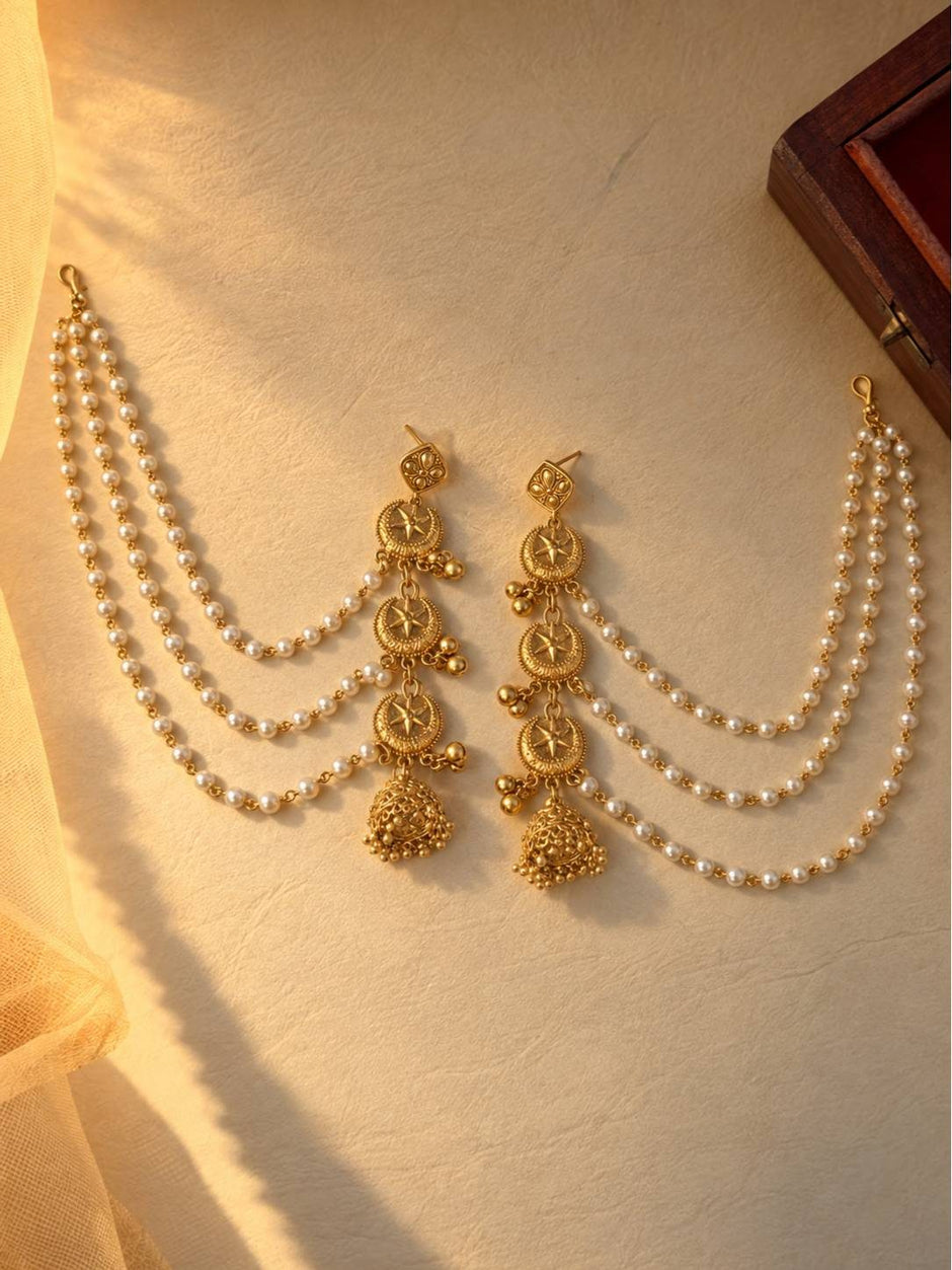Pair of gold-plated jhumka earrings with three-strand faux pearl hair chains laid flat on a cream surface