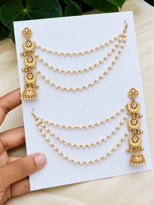 Both gold-plated pearl hair chain jhumka earrings displayed on a white card held by a hand against green leaves