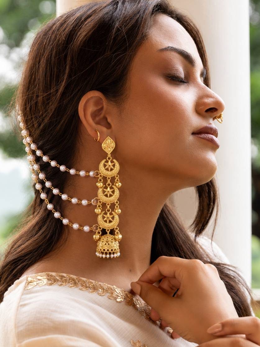 Woman wearing a single gold-plated crescent jhumka with pearl hair chain draped over her ear in a white outfit