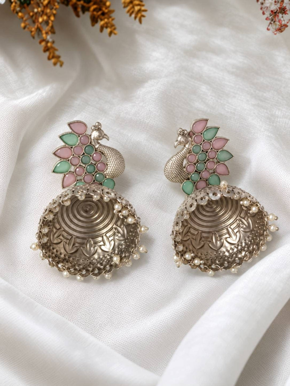 Two oxidised silver peacock jhumka earrings laid flat on white fabric showing engraved dome and pearl fringe