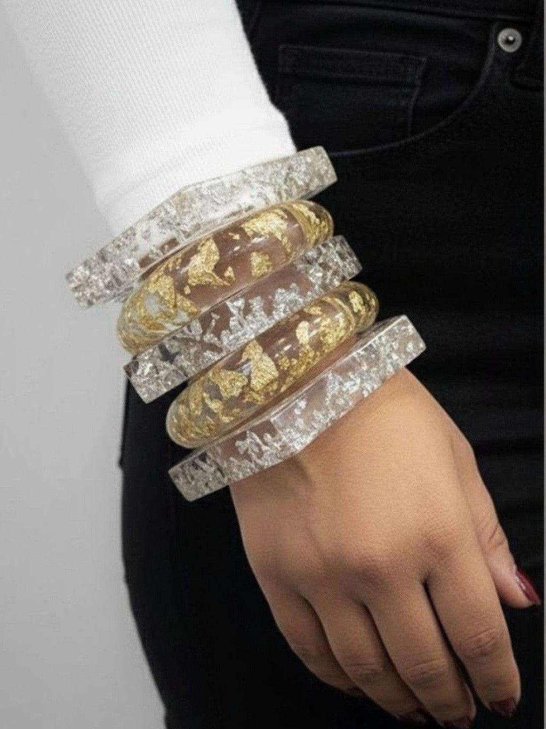 Five clear resin bangles stacked on a woman's wrist showing gold and silver foil inclusions against a white sleeve and black jeans