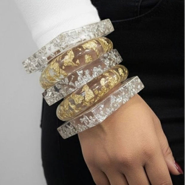 Five clear resin bangles stacked on a woman's wrist showing gold and silver foil inclusions against a white sleeve and black jeans