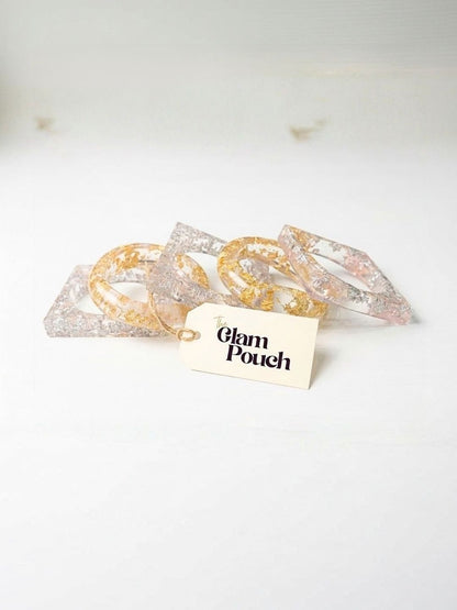 Five clear resin bangles with gold and silver foil arranged loosely on a white surface beside a Glam Pouch branded tag