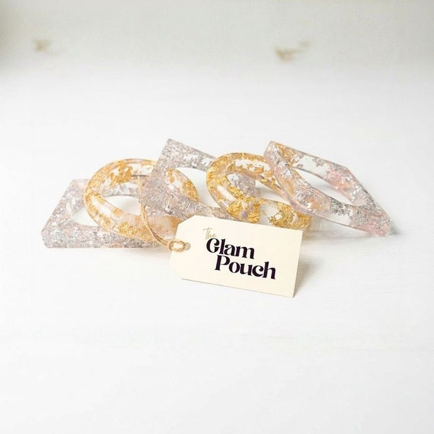 Five clear resin bangles with gold and silver foil arranged loosely on a white surface beside a Glam Pouch branded tag