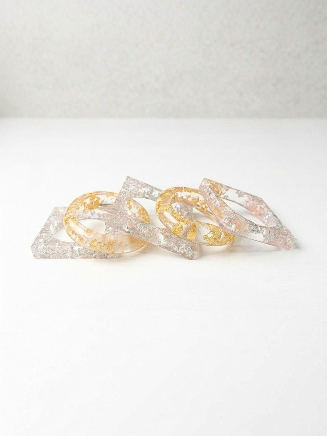 Close-up of five clear resin bangles in square and oval silhouettes showing embedded metallic foil on a white background