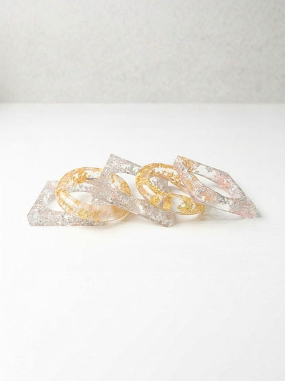 Close-up of five clear resin bangles in square and oval silhouettes showing embedded metallic foil on a white background