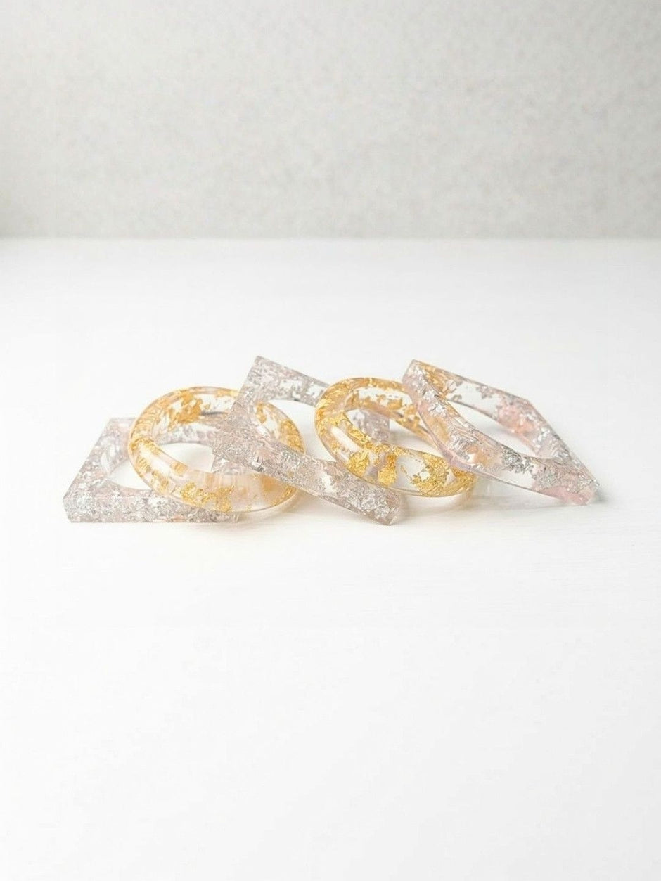 Close-up of five clear resin bangles in square and oval silhouettes showing embedded metallic foil on a white background