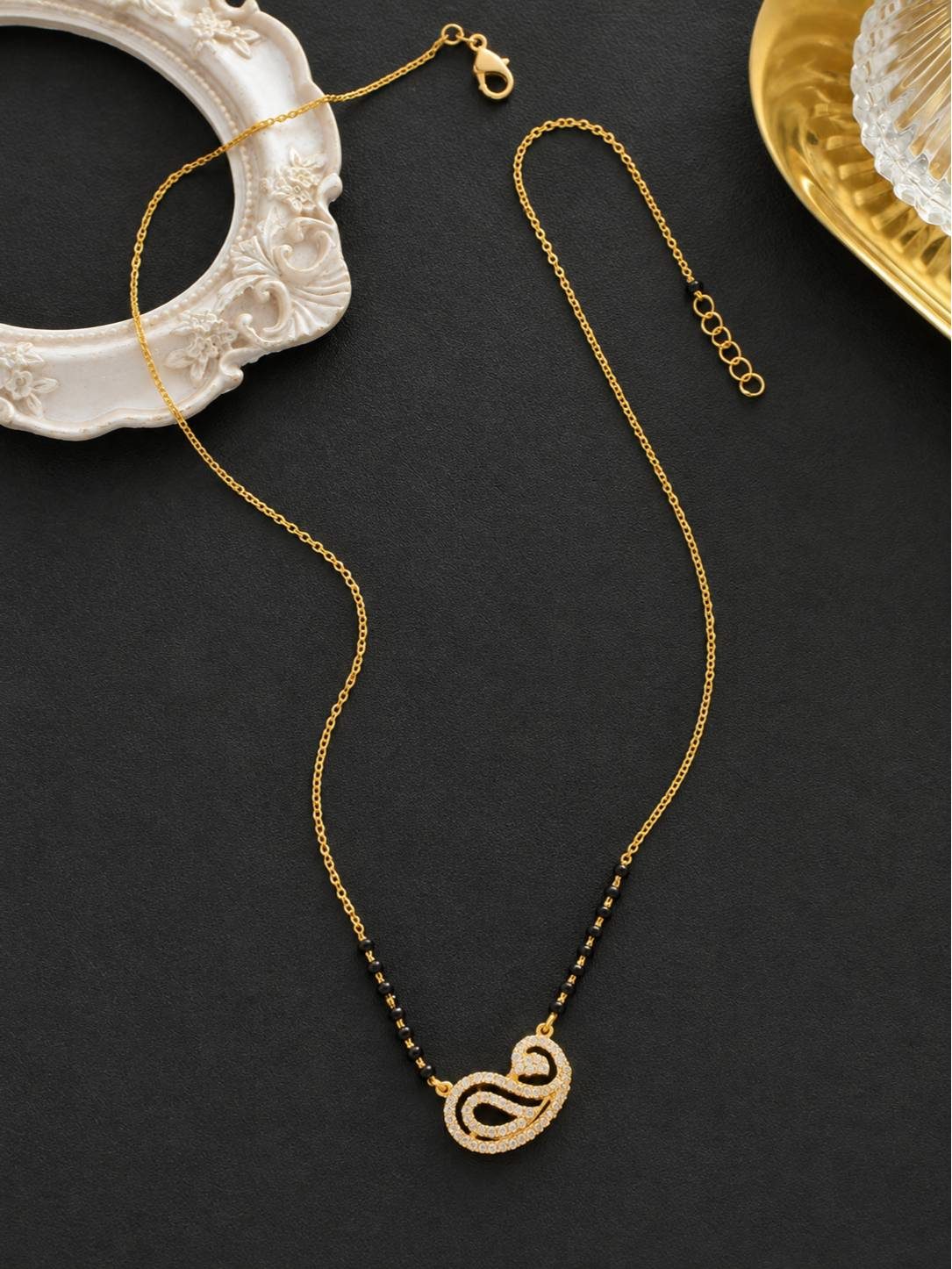 Gold-plated CZ mangalsutra laid flat on black surface beside decorative white frame showing lobster clasp