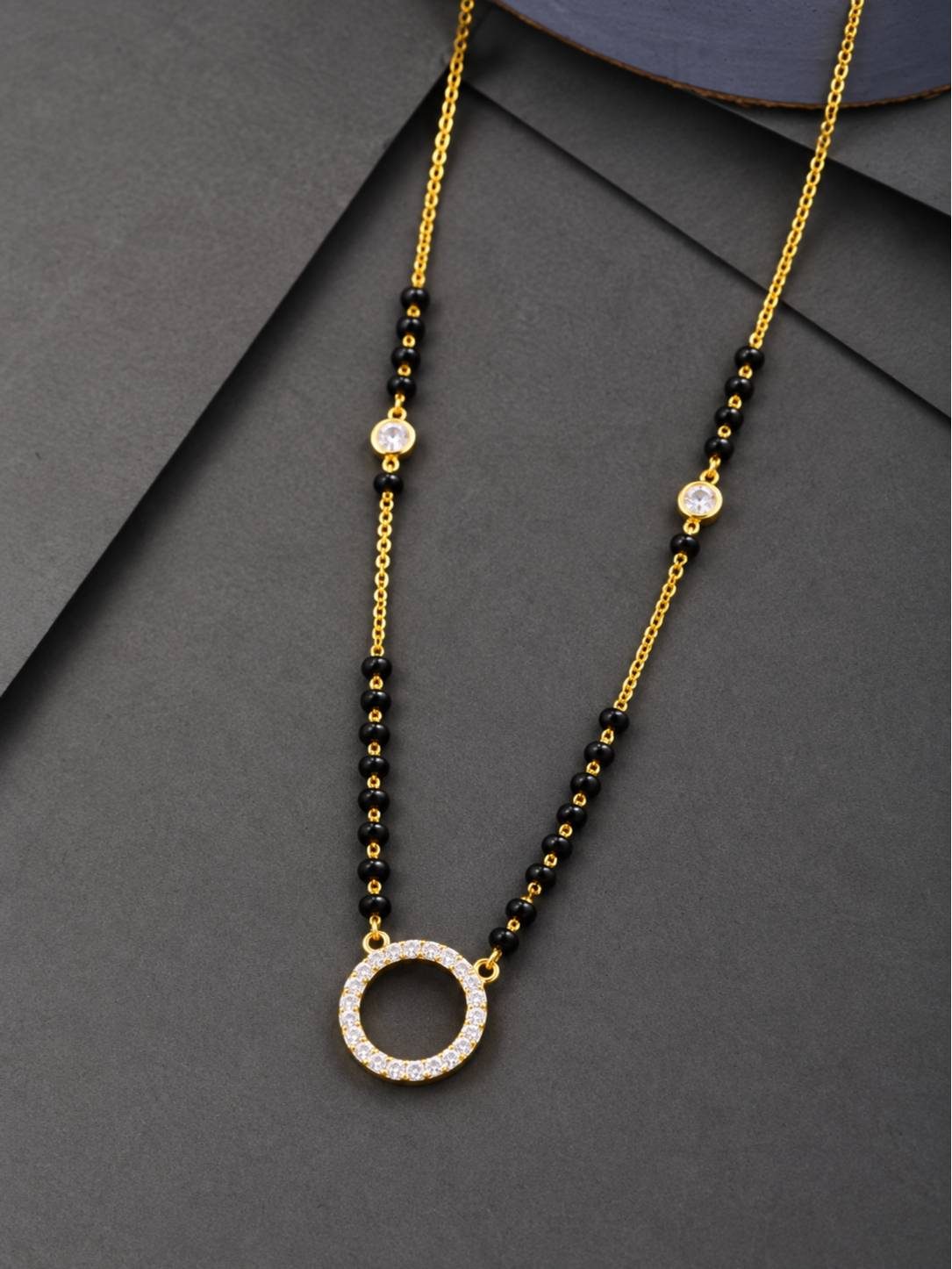 Gold-plated black bead mangalsutra with open circle CZ pendant laid flat on dark grey surface