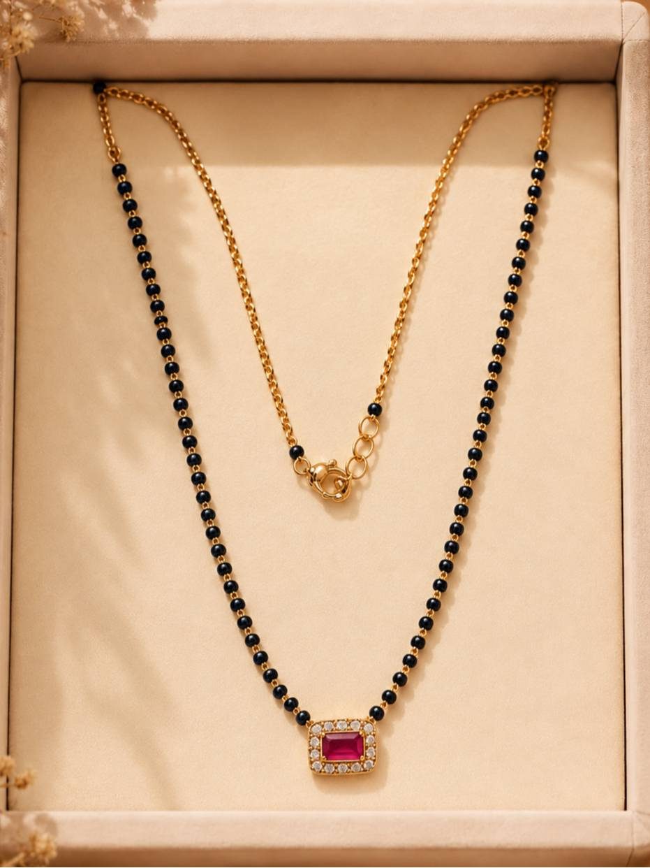 Gold-plated black bead mangalsutra with fuchsia CZ square pendant displayed flat in a cream jewellery box