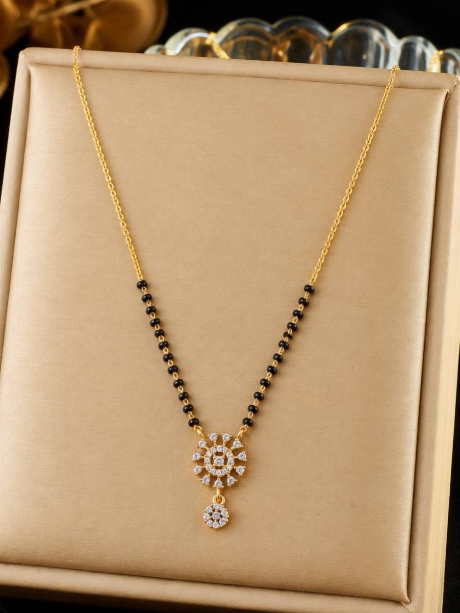 Gold-plated CZ mangalsutra with black bead strands and sunburst pendant displayed on a beige jewellery box
