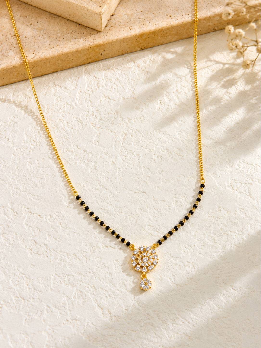 CZ mangalsutra with black beads and gold chain laid flat on a textured white surface with dried flowers