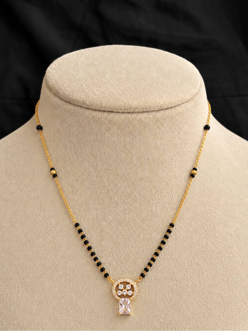 Gold-plated mangalsutra with black beads and circular CZ pendant displayed on a beige jewellery bust