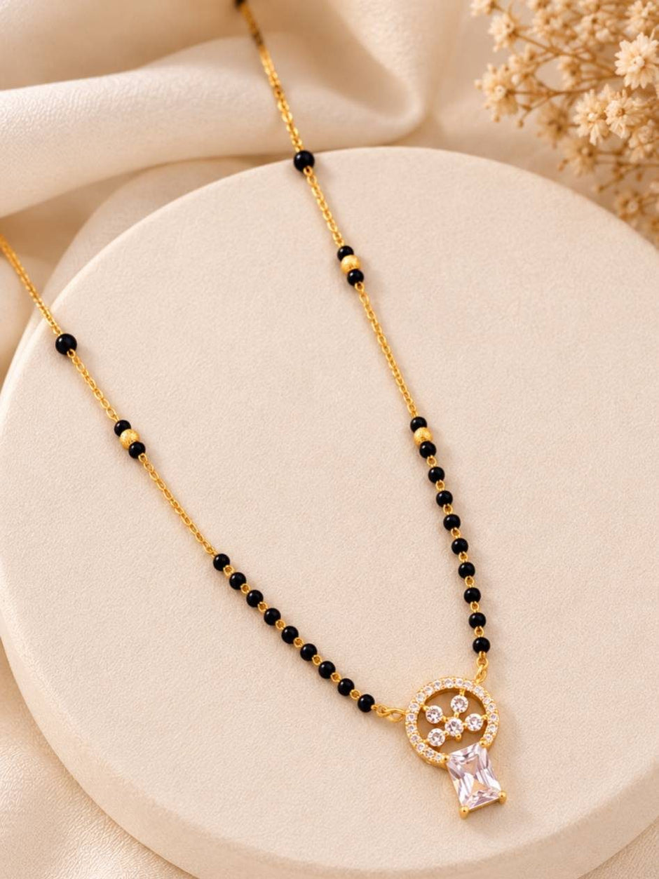 Gold-plated black bead mangalsutra with CZ circular pendant and baguette stone drop laid on a round cream surface
