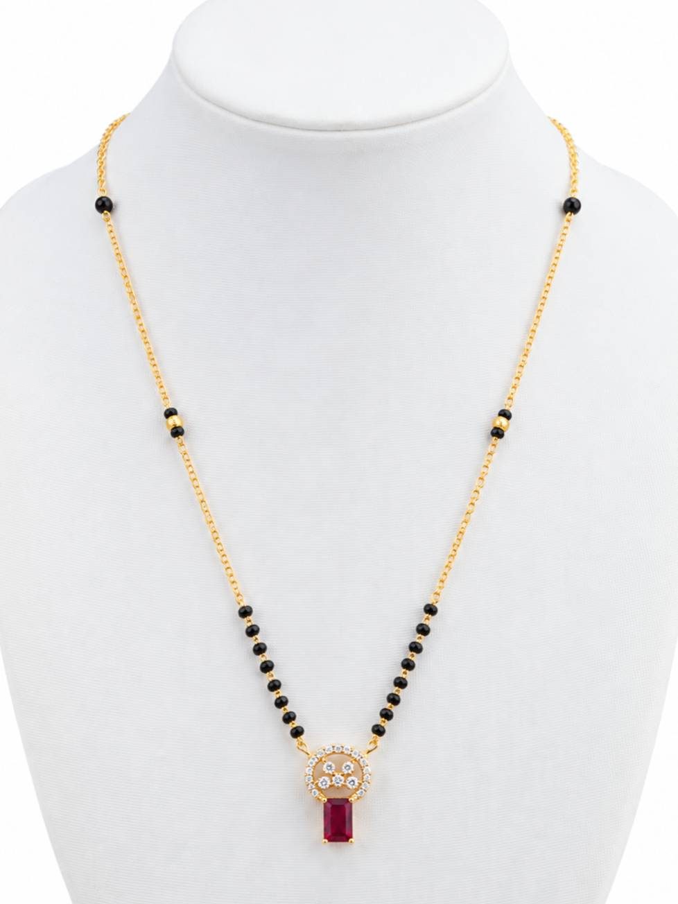 Gold-plated black bead mangalsutra with circular CZ and ruby-red pendant displayed on white jewellery bust