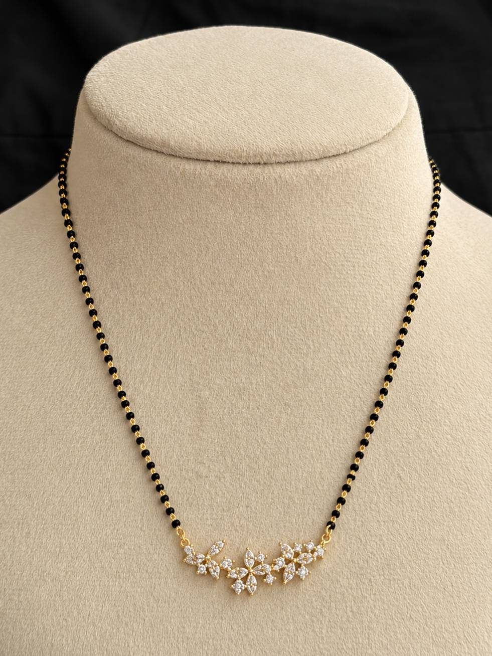 Gold-plated CZ floral mangalsutra with black beads displayed on a beige jewellery bust