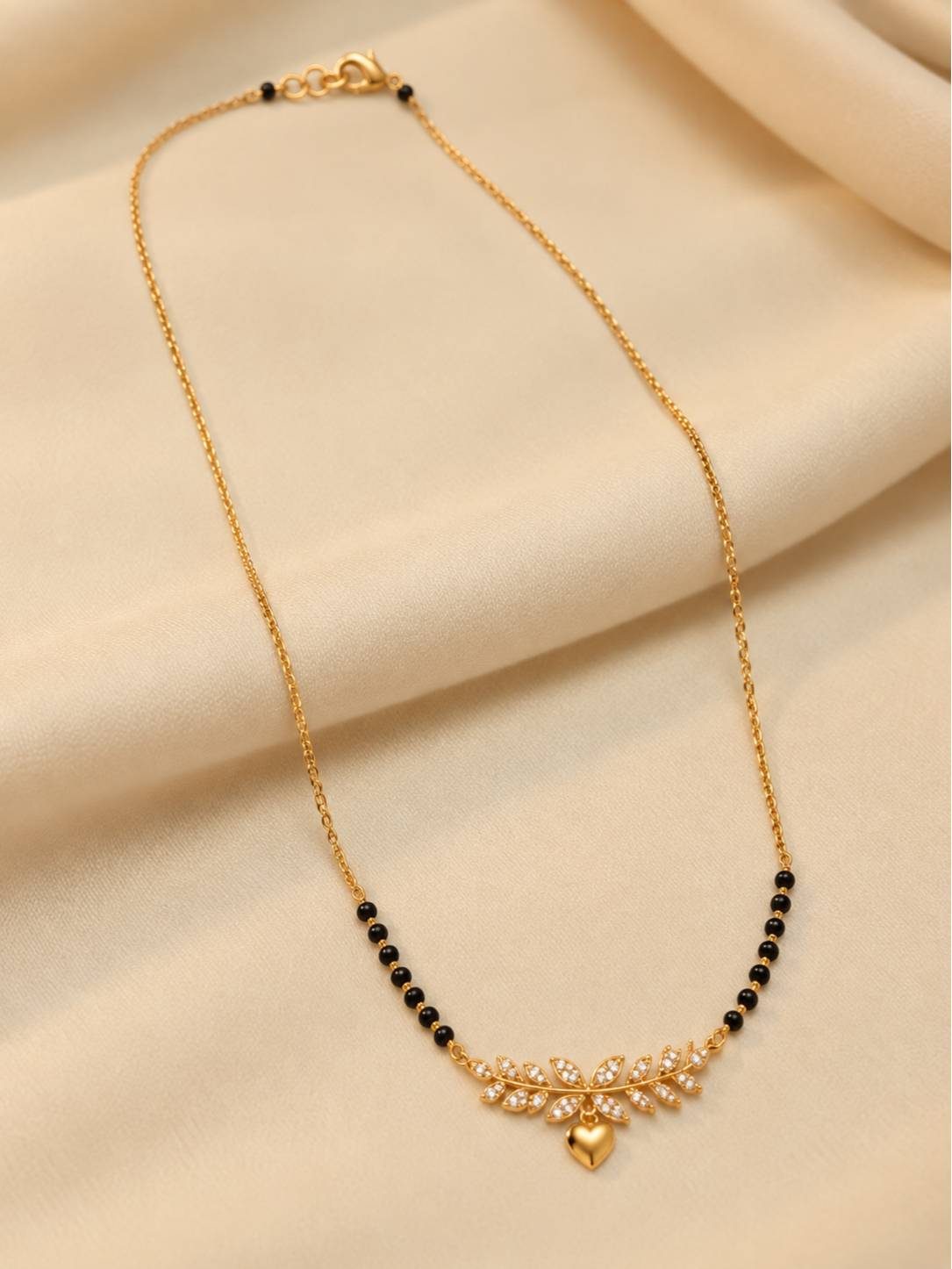 Full-length view of gold-plated black bead mangalsutra with leaf and heart pendant on cream fabric