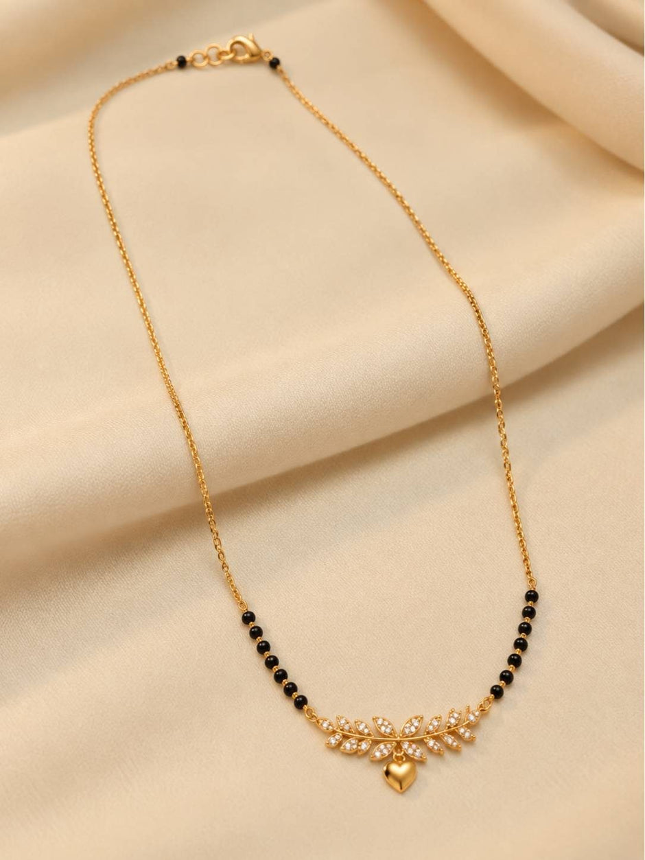 Full-length view of gold-plated black bead mangalsutra with leaf and heart pendant on cream fabric