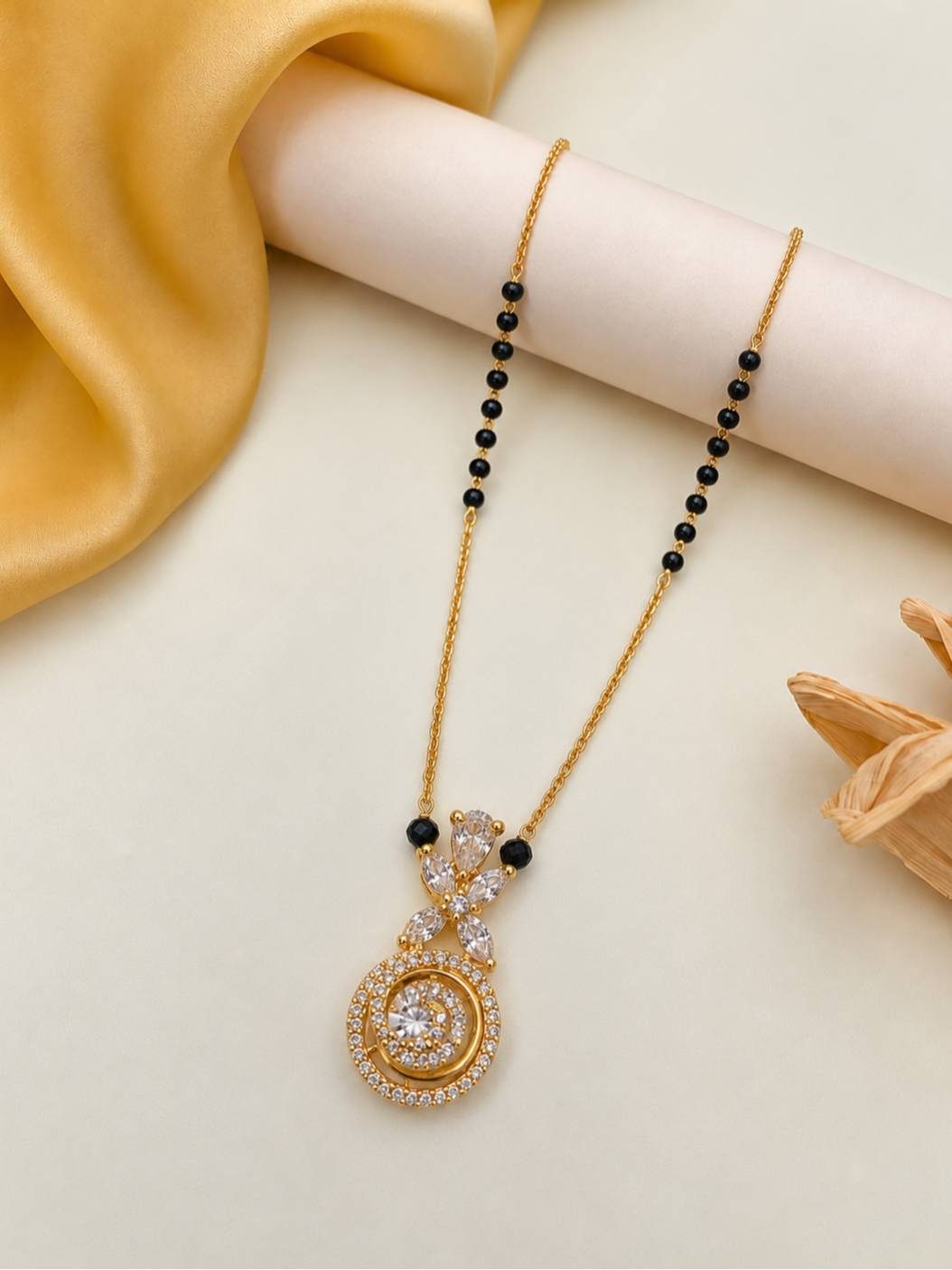 Gold-plated mangalsutra with circular CZ pendant laid on a cream surface beside dried flowers