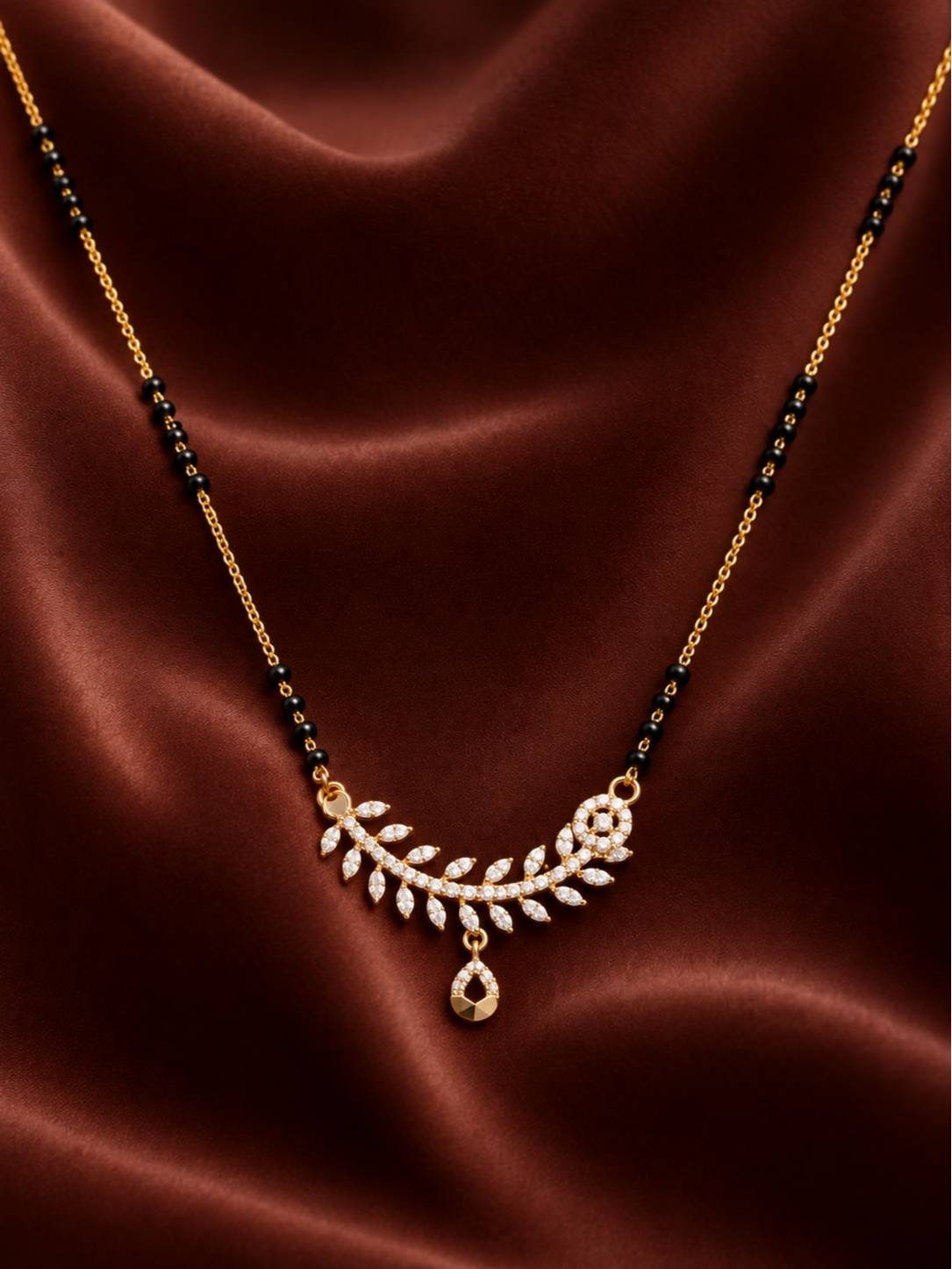 Close-up flat-lay of the curved wheat-leaf CZ pendant and black bead chain on deep red satin fabric