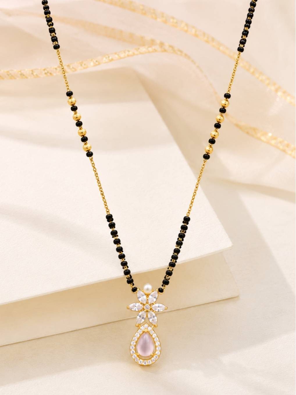 Gold-plated mangalsutra laid flat on a white surface with golden ribbon accent, highlighting the full length of the black bead chain and CZ pendant