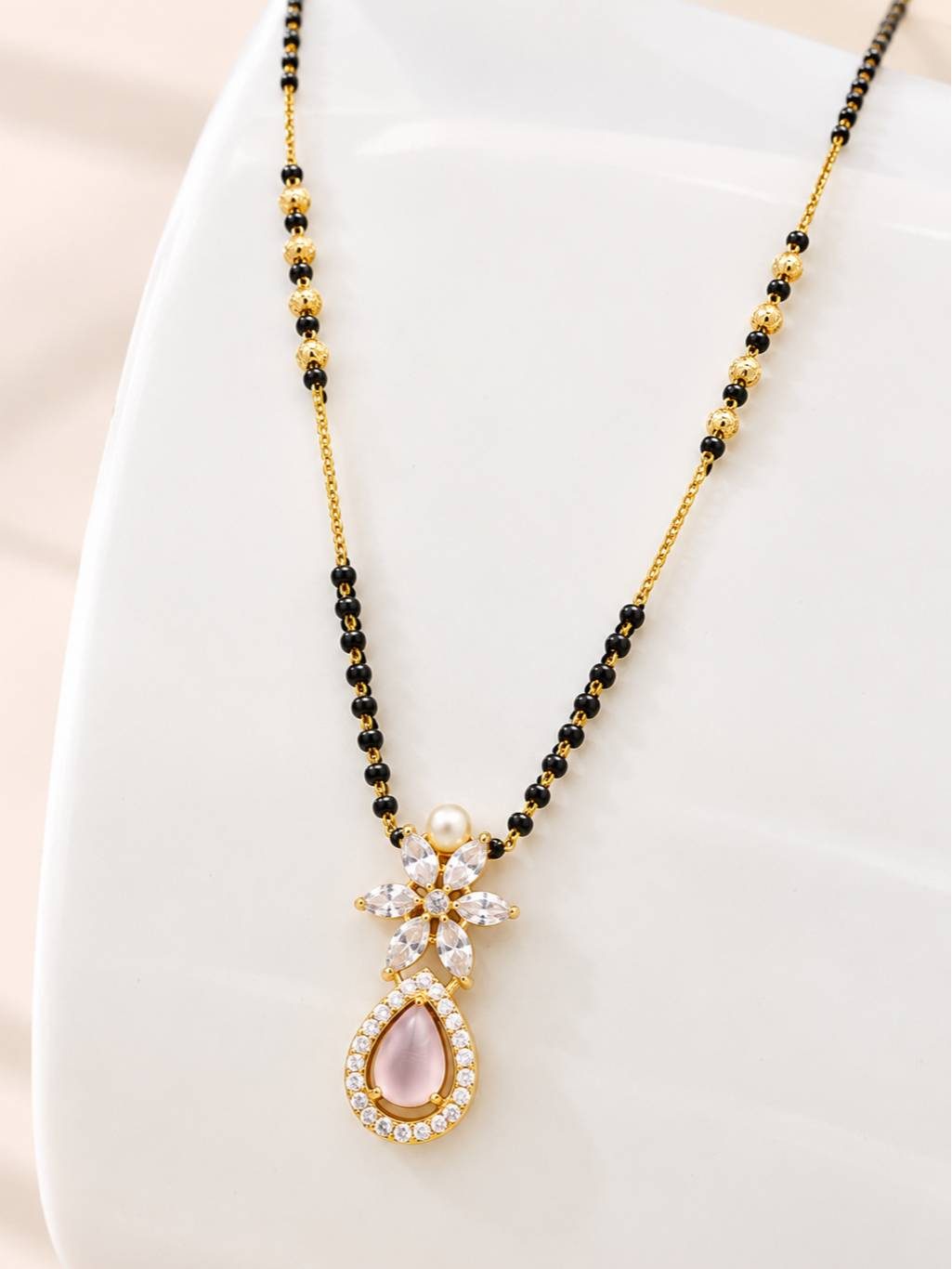 Close-up of mangalsutra pendant on a white curved display stand showing marquise CZ flower, pearl, and rose cabochon teardrop in gold frame