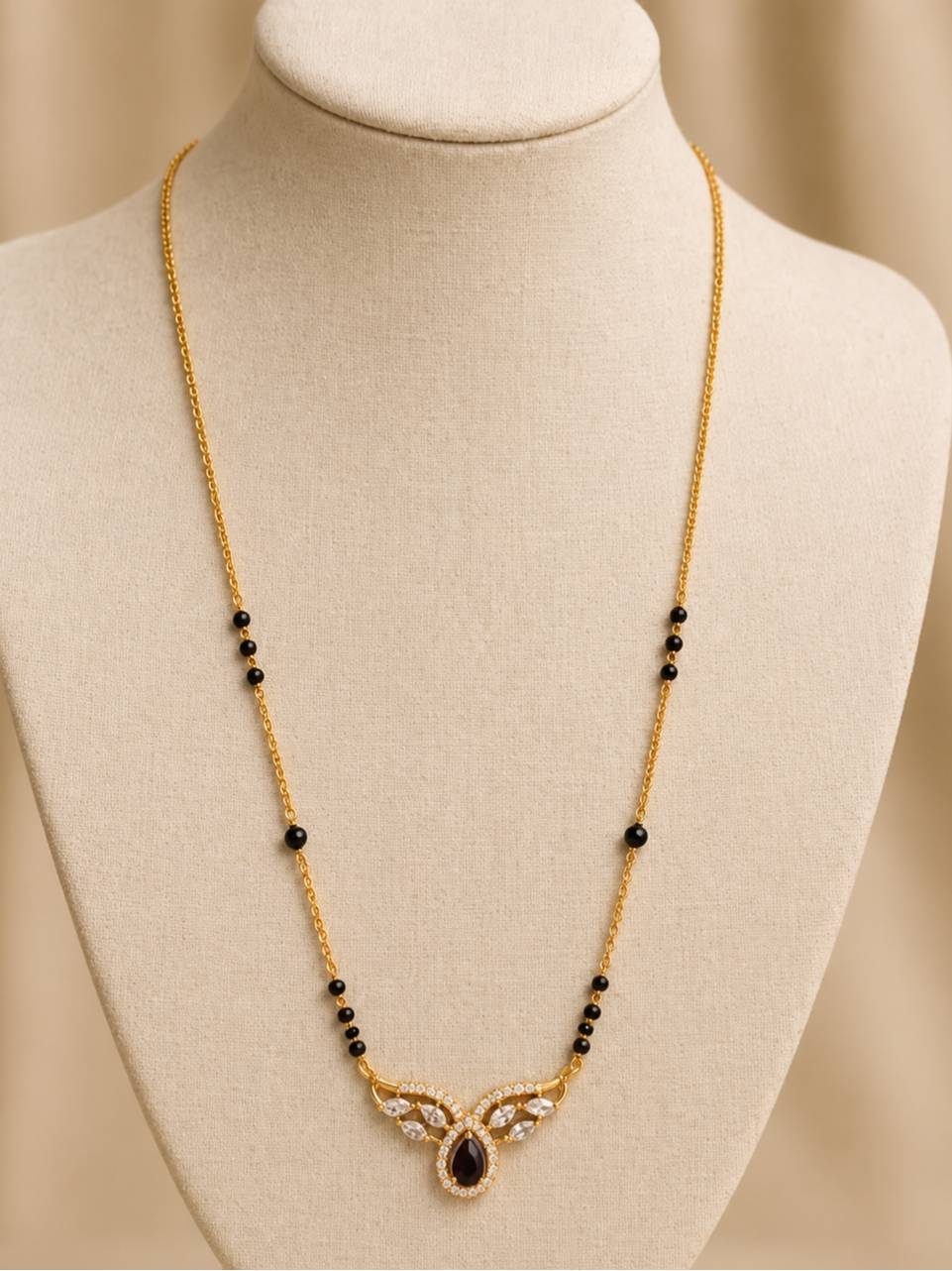 Gold-plated black bead mangalsutra displayed on a cream jewellery bust with CZ and red teardrop pendant